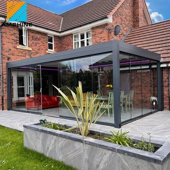 Aluminium Pergola NSW 4 A sleek dark aluminium pergola encloses a brick home patio, with glass panels, purple lighting and red seating beside a landscaped planter.